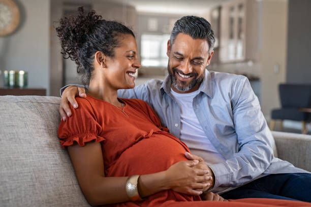 Mid adult couple expecting a baby while husband caresses the belly of his pregnant african woman. Happy father hands on expecting mother's baby bump while embracing her on couch. Lovely indian man touching belly of his girlfriend and feeling baby mov